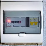 Cyfrowy licznik prądu w białej obudowie, obok dwa wyłączniki. Zastanawiasz się, ile kosztuje sprawdzenie licznika prądu?