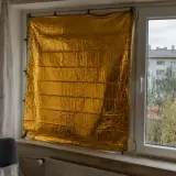 Złoty koc termiczny na okno, chroniący przed zimnem. W tle widać budynek mieszkalny i drzewa.