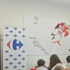 Mężczyzna w garniturze wręcza książkę "Karta Dużej Rodziny" na tle banerów Energa i Carrefour.