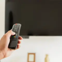 Dłoń trzymająca pilot do telewizora na tle wyłączonego ekranu.