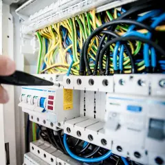 Dłoń z narzędziem przy skrzynce z bezpiecznikami i kolorowymi przewodami elektrycznymi.