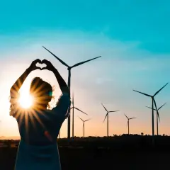 Sylwetka kobiety tworzy serce na tle zachodzącego słońca i farmy wiatrowej. Miłość do czystych źródeł energii.
