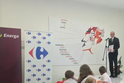 Mężczyzna w garniturze wręcza książkę "Karta Dużej Rodziny" na tle banerów Energa i Carrefour.