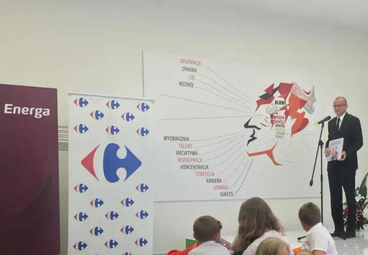 Mężczyzna w garniturze wręcza książkę "Karta Dużej Rodziny" na tle banerów Energa i Carrefour.
