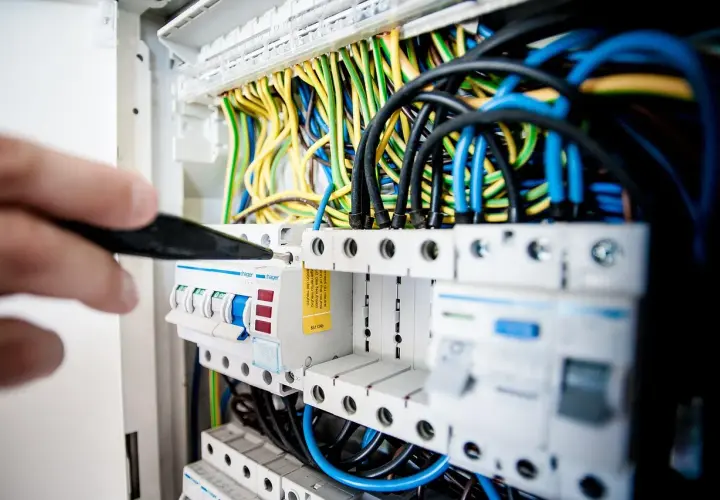 Dłoń z narzędziem przy skrzynce z bezpiecznikami i kolorowymi przewodami elektrycznymi.