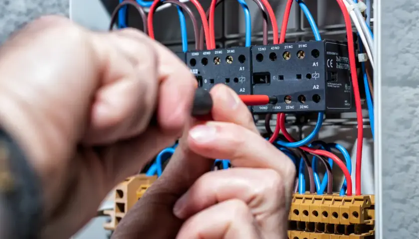 Ręce elektryka pracują przy rozdzielnicy, podłączając przewody. Widać styki i bezpieczniki, a napięcie w sieci jest pod kontrolą.