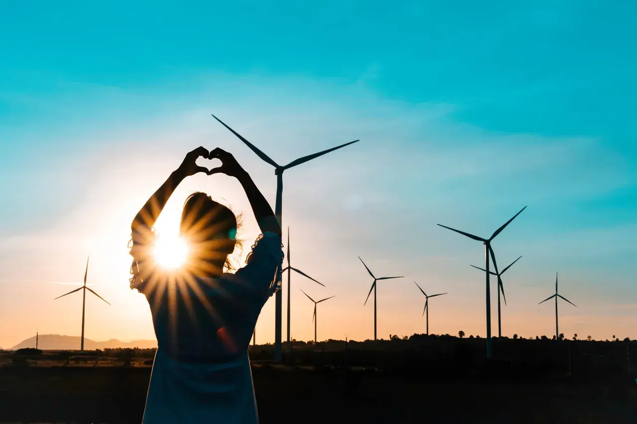 Sylwetka kobiety tworzy serce na tle zachodzącego słońca i farmy wiatrowej. Miłość do czystych źródeł energii.
