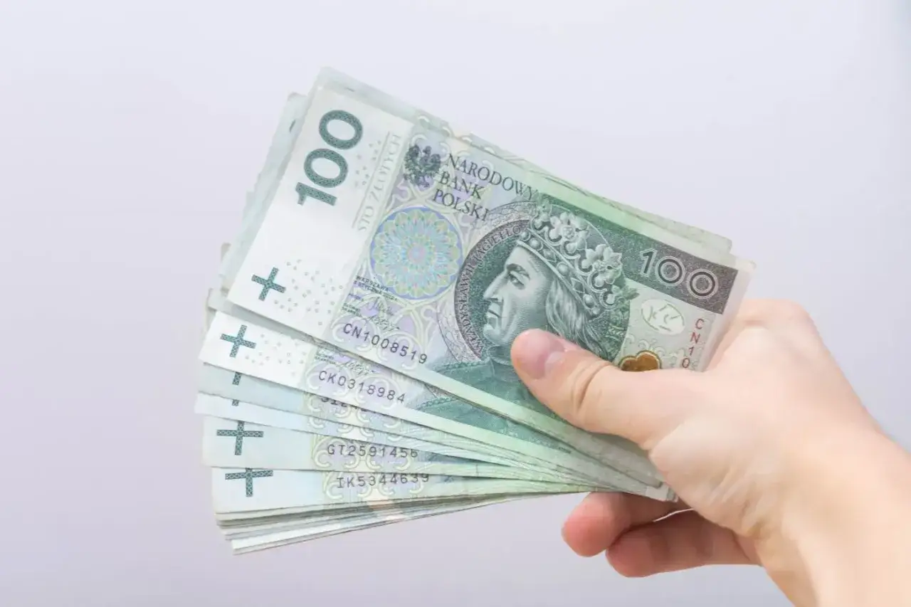 Dłoń trzyma stos banknotów 100 zł. Może to być pomoc w opłaceniu rachunków, np. bon energetyczny 2026.